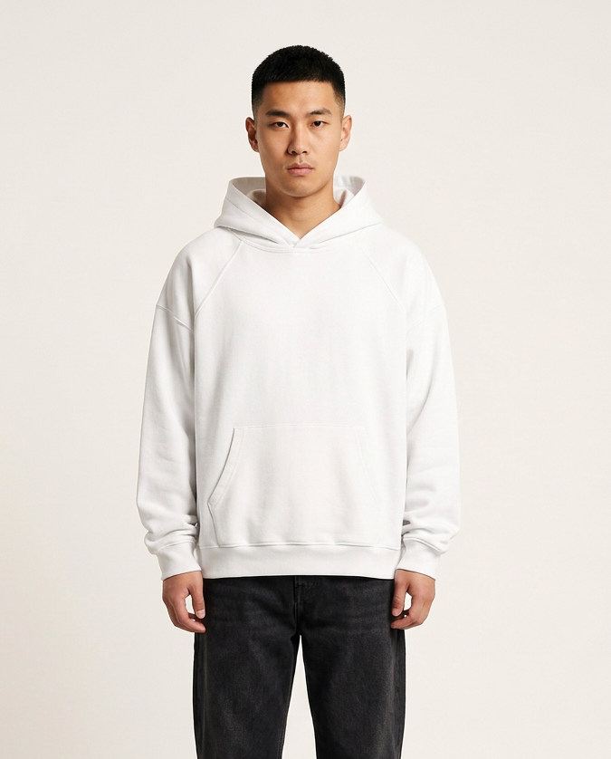 A man wearing a plain white pullover hoodie with black jeans and black shoes, standing against a solid off-white studio background.